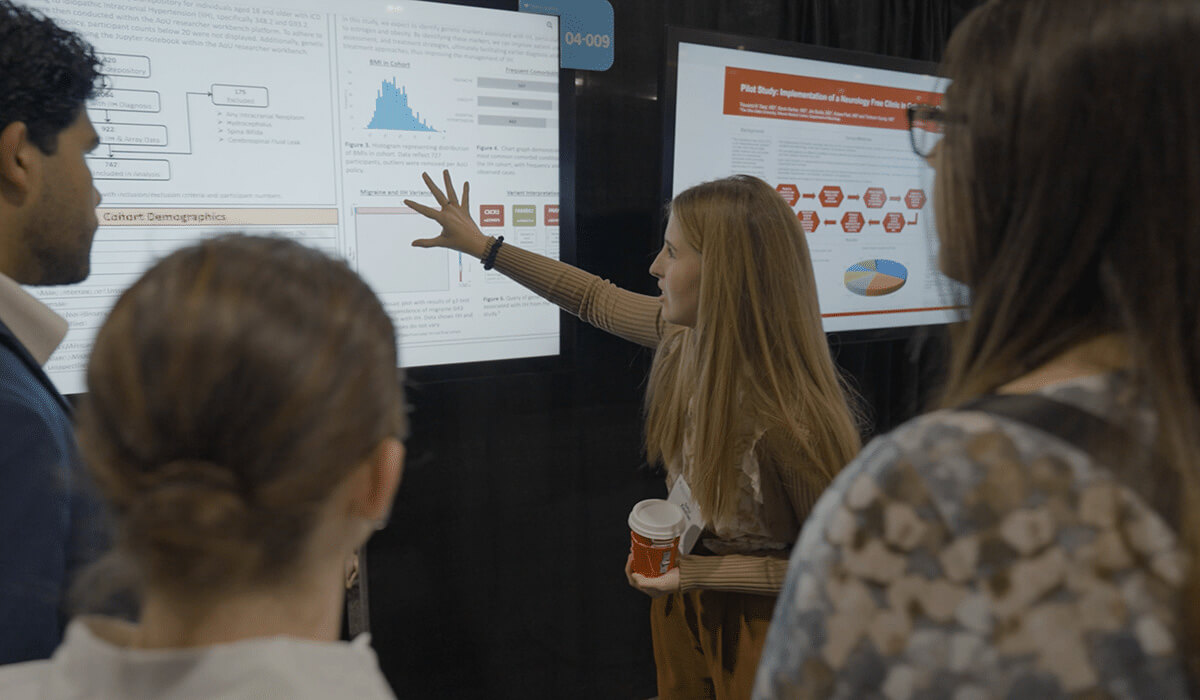 A woman points to data on a large screen while explaining to three people. The group stands in front of scientific posters and presentations at what appears to be a conference or academic event.